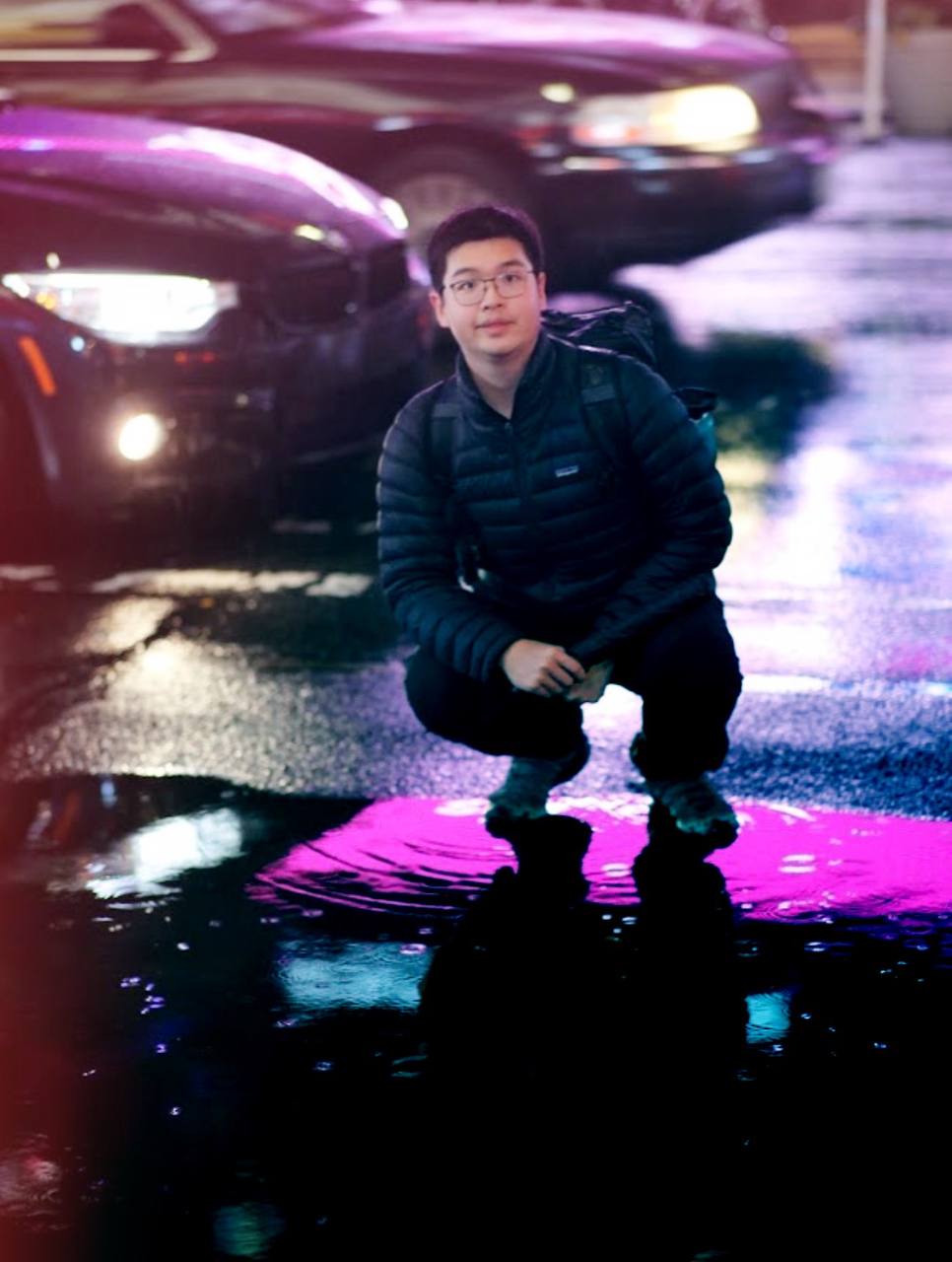 Jack crouching in a neon-lit rainy parking lot, pink puddle reflection